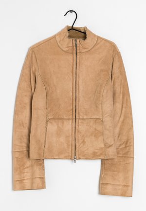 Beige mokkanahkatakki, jossa edessä vetoketju, korkea kaulus, pitkät hihat ja saumadetaljit, roikkuu mustassa henkariassa valkoista taustaa vasten.