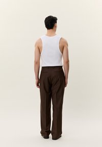 Moški, ki stoji obrnjен stran, oblečen v belo brezrokavnik in rjave po meri krojene hlače na enostavni svetli podlagi.