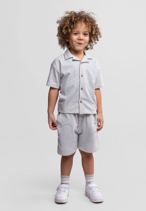 Małe dziecko z kręconymi włosami uśmiecha się, nosząc krótką koszulę na guziki i pasujące paskowane spodenki oraz białe trampki i skarpetki.