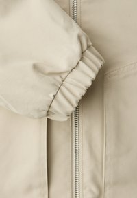 Manica di giacca beige con polsino elastico accanto a una cerniera verticale argento e a una tasca rettangolare cucita su uno sfondo di tessuto liscio.