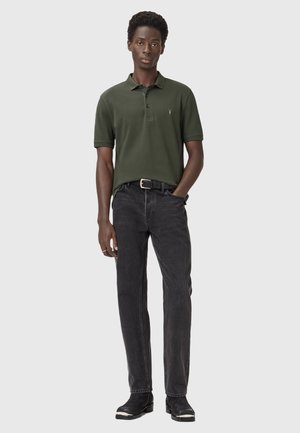 Olivgrön polo med tre knappar, svarta jeans och svarta ankelstövlar. Polon har en liten, diskret logotyp på bröstet.