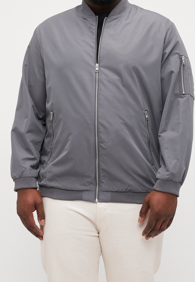 Homme portant une veste bomber gris zippée avec deux poches avant et une poche zippée sur la manche gauche, associée à un pantalon beige clair.