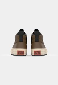 Brown high-top sneakers with black accents, textured material, and a cream-colored sole. Features red branded logo on the heel.