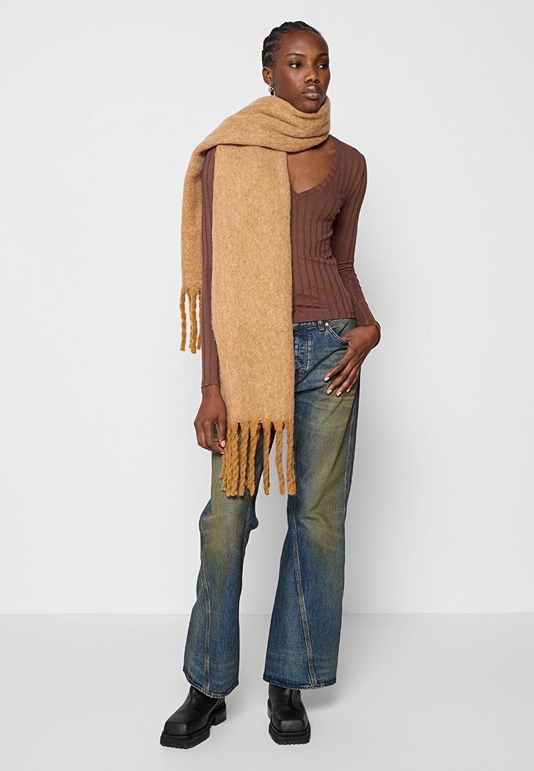 Écharpe en laine couleur camel avec des franges, portée sur un haut brun à côtes et assortie à un jean à jambes larges et des bottines noires.