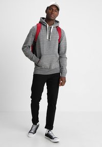 Sudadera con capucha gris y bolsillo tipo canguro, combinada con jeans negros y zapatillas blancas. La persona lleva una mochila roja y usa una gorra.