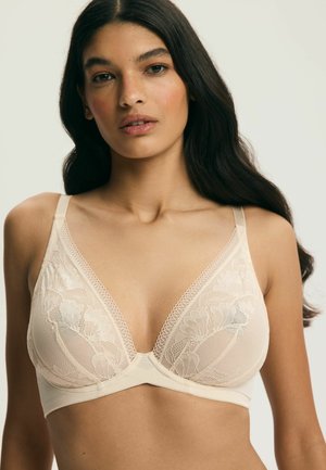 Žena s dlhými tmavými vlasmi nosiaca béžovú čipkovanú podprsenku s kvetinovými vzormi a nastaviteľnými ramienkami na jednoduchom svetlom pozadí.