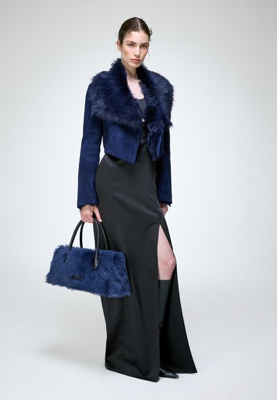 Femme portant une veste bleu marine à col en fourrure et une robe noire fendue à la cuisse, tenant un sac à main en fourrure bleu marine assorti.