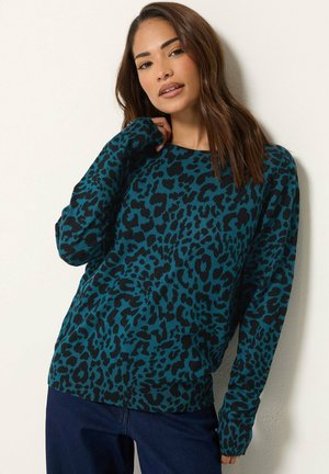 Femme portant un pull à manches longues imprimé léopard vert d'eau et noir et un jean bleu foncé, debout devant un fond clair uni.