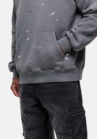 Felpa grigia con design a spruzzi di vernice rosa e bianca, dotata di tasca frontale e polsini a costine, indossata con pantaloni cargo scuri.