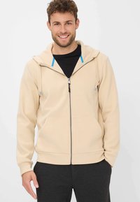 Felpa zip-up beige con una consistenza morbida, cordini regolabili e accenti blu a contrasto sul cappuccio; abbinata a pantaloni scuri.