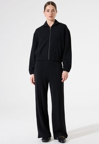Zwarte cropped zip-up jas met hoge kraag en rib gebreide manchetten, gecombineerd met losvallende zwarte broeken met verticale naden en een gladde textuur.