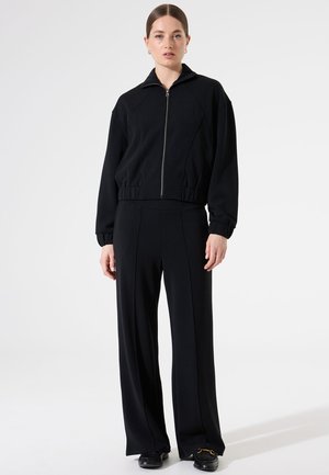 Veste noire cropped avec fermeture éclair, col montant et poignets côtelés, associée à un pantalon noir ample avec des coutures verticales et une texture lisse.