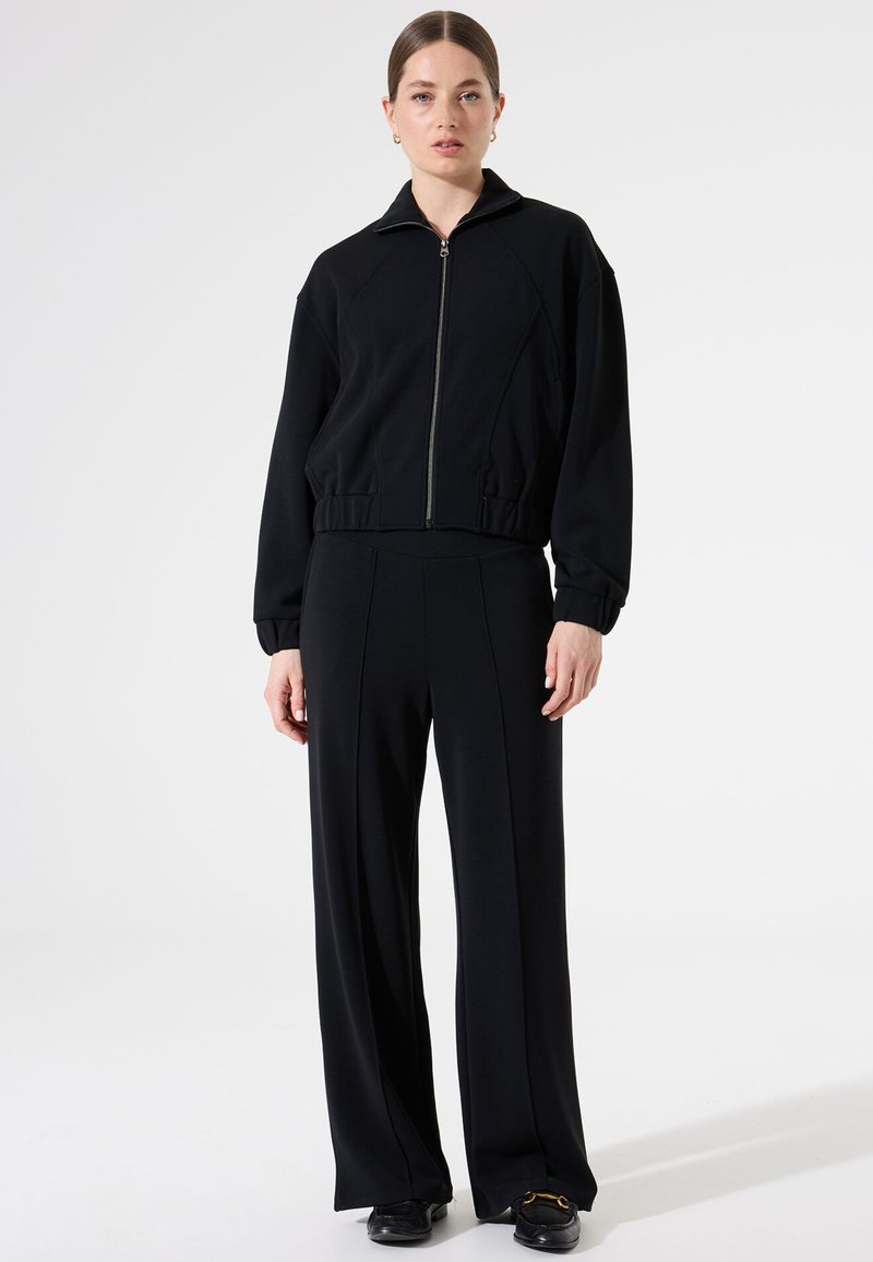 Zwarte cropped zip-up jas met hoge kraag en rib gebreide manchetten, gecombineerd met losvallende zwarte broeken met verticale naden en een gladde textuur.