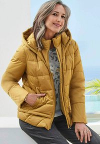 Senfgelb gepolsterte Jacke mit Kapuze, gestepptem Design, Reißverschluss und Seitentaschen. Getragen über einem gemusterten grauen Pullover.