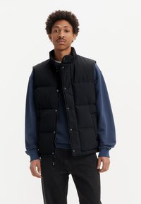 Svart vadderad väst med hög krage, tryckknappar och sidofickor, bärs över en blå sweatshirt och svarta byxor. Slät textur.