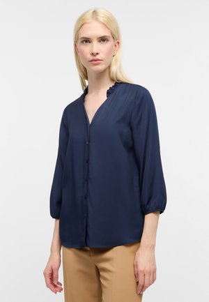 Marineblaue Bluse aus glattem Stoff mit V-Ausschnitt, Rüschensaum und dreiviertellangen Puffärmeln; Knopfverschluss an der Vorderseite.