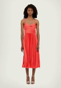 Rūpīgi salocīta koralaina kleita ar plāniem siksniņām, ar mezglu pie krūšdaļas un plūstošu svārku. Valkāta ar baltām kurpēm un rakstainu somu.