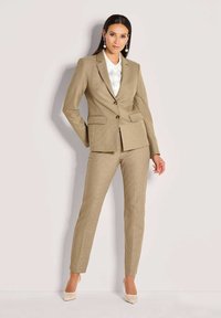 Beige op maat gemaakt pak met een gestructureerde blazer en aansluitende broek. De blazer heeft twee voorzakken, een enkele knoopsluiting en een wit overhemd eronder.