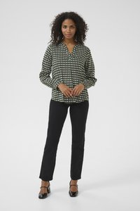 Woman with curly hair wearing a green and white patterned blouse, black pants, and black heels standing against a plain background.