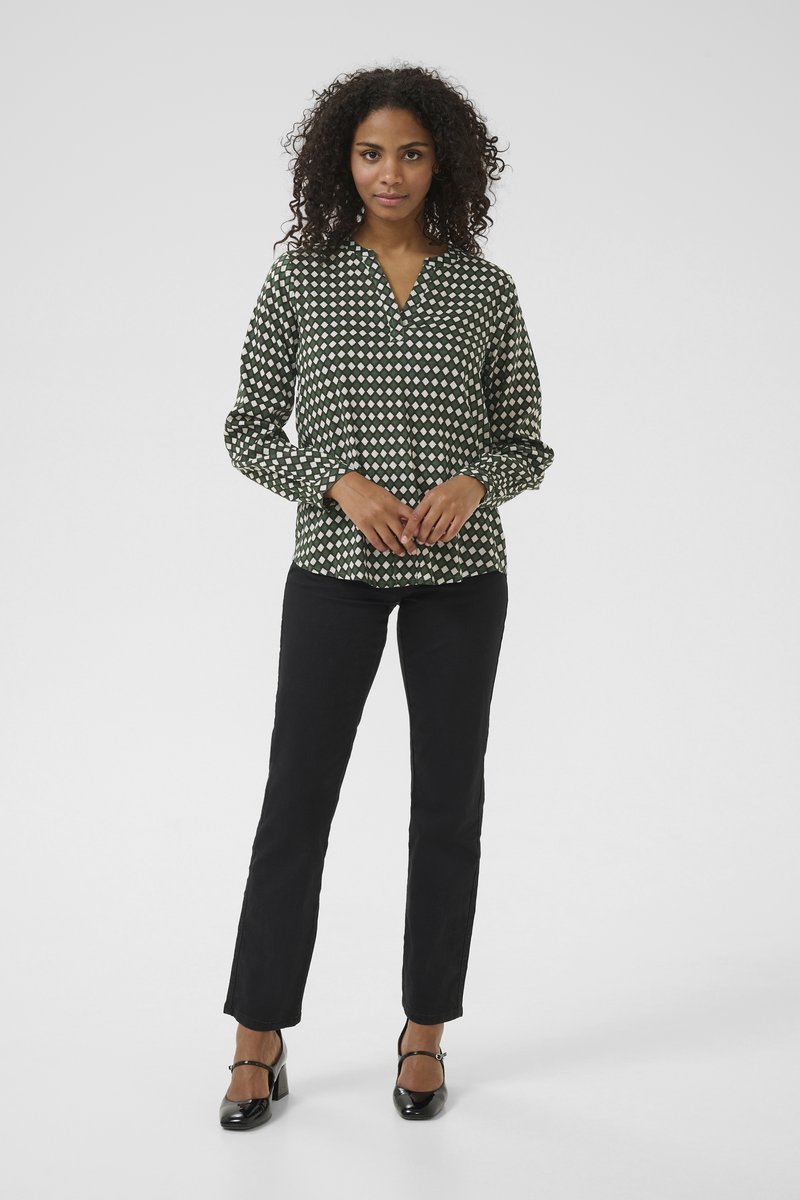 Woman with curly hair wearing a green and white patterned blouse, black pants, and black heels standing against a plain background.