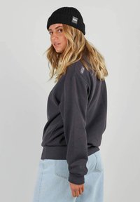 Sweatshirt gris foncé à manches longues, avec des poignets côtelés et un ourlet ajusté. Associé à un bonnet en tricot noir portant une étiquette en patch. Jupe en denim clair.