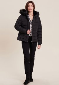 Donna che indossa una giacca nera trapuntata con cappuccio in pelliccia, una blusa con fantasia, pantaloni neri e stivaletti neri, in piedi davanti a uno sfondo chiaro.