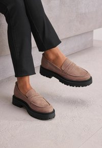 Mocassini in suede rosa con suola in gomma nera spessa, caratterizzati da dettagli cuciti e una superficie liscia. Indossati con pantaloni neri alla caviglia.