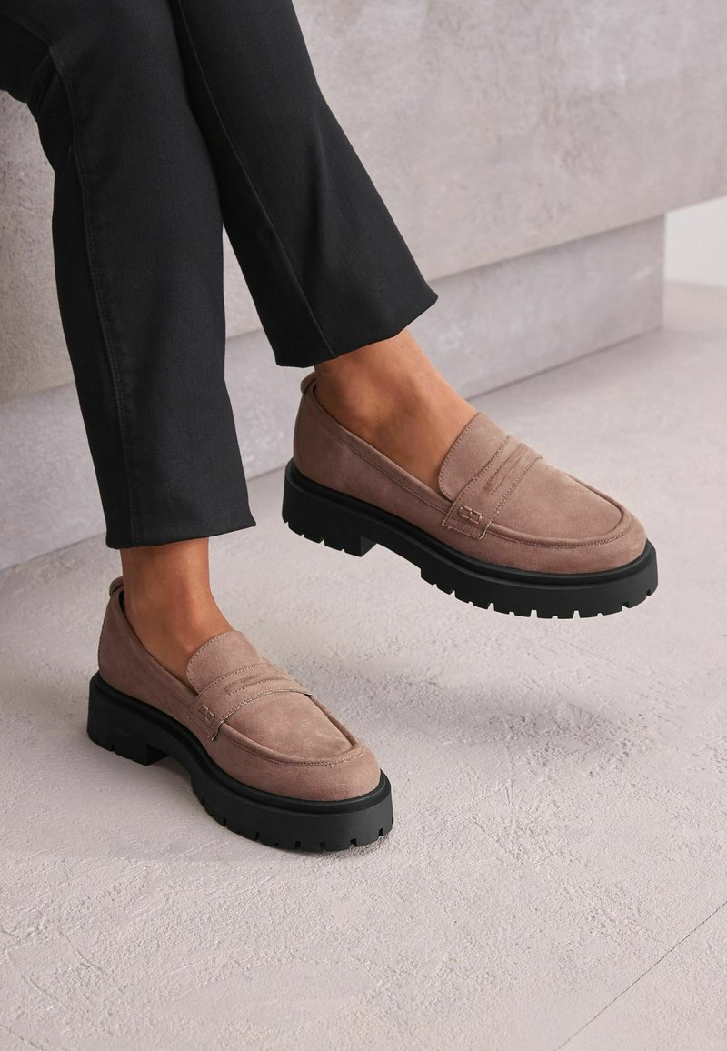 Mocassini in suede rosa con suola in gomma nera spessa, caratterizzati da dettagli cuciti e una superficie liscia. Indossati con pantaloni neri alla caviglia.