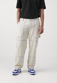 Pantalones de mezclilla color beige claro con costura diagonal, corte relajado y un bolsillo tipo parche. Combinados con zapatillas azul y blanco.