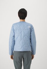 Jasnoniebieska pikowana kurtka z miękkiego materiału, z okrągłym dekoltem i logo na ramieniu, zestawiona z szarymi spodniami.