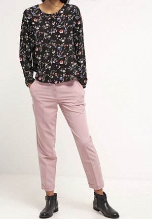 Nainen pukeutuneena mustaan kukalliseen pitkähihainen puseroon, vaaleanpunaisiin kapeneviin housuihin ja mustiin nilkkureihin, seisomassa käsivarret taskuissa.