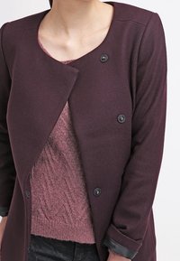 Burgundská vlněná směsová bunda s kulatým výstřihem, asymetrickým zapínáním vpředu a dvěma černými knoflíky. Pod ní je měkký růžový pletený svetr.