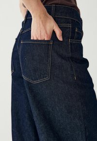 Sötétkék farmernadrág hátsó zsebbel, amelyen látható a varrás. Az anyag textúrája kidolgozott, a dizájn pedig magasan szabott derékkal rendelkezik.