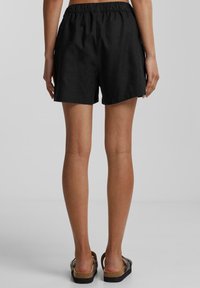 Femme portant un short noir à taille élastique et des sandales marron, se tenant de dos sur un fond gris uni.