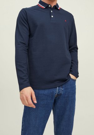 Mand iført en navyblå polotrøje med lange ærmer med rødt og hvidt kantbånd ved kraven og blå jeans, stående med den ene hånd i lommen.