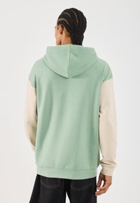 Uomo con capelli intrecciati che indossa un hoodie verde con maniche beige, in piedi di spalle contro uno sfondo bianco semplice.