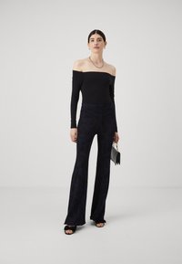 Zwarte off-shoulder top met lange mouwen, gecombineerd met hooggesneden, flared donker patroon pantalons. Het model houdt een kleine zwarte handtas vast.