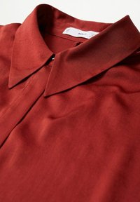 Blouse couleur rouille avec une texture lisse, dotée d'un col classique et d'une fermeture à boutons au niveau du cou. Le tissu présente un drapé subtil.