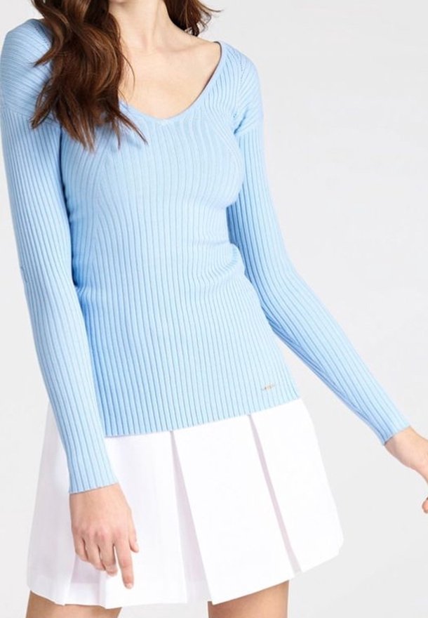 Femme portant un haut côtelé bleu clair à manches longues et une jupe plissée blanche sur un fond uni.