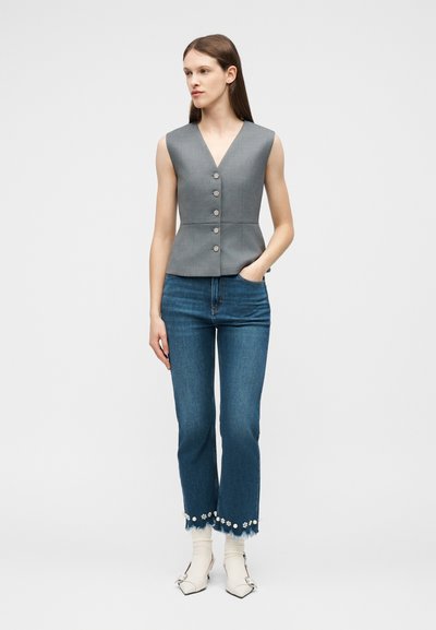 Γυναίκα που φοράει γκρι αμάνικο γιλέκο με κουμπιά, μπλε τζιν με φλοράλ διακόσμηση, λευκές κάλτσες και μυτερές λευκές μπότες, στέκεται μπροστά σε απλό φόντο.