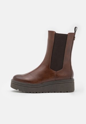 Botte Chelsea en cuir marron avec semelle noire épaisse et rainurée, panneau latéral élastique et languette à l'arrière sur fond blanc.