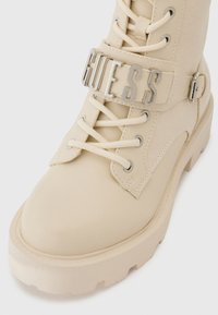 Bottes chelsea beiges à lacets avec une finition lisse, présentant un panneau latéral texturé et une bande logo en argent bien visible autour de la cheville.