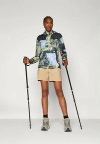 Pohodniški komplet, ki vključuje potiskan, lahek dežni plašč v modrih in zelenih odtenkih, kratke hlače in pohodne palice. Čevlji so sivi z črnimi detajli.