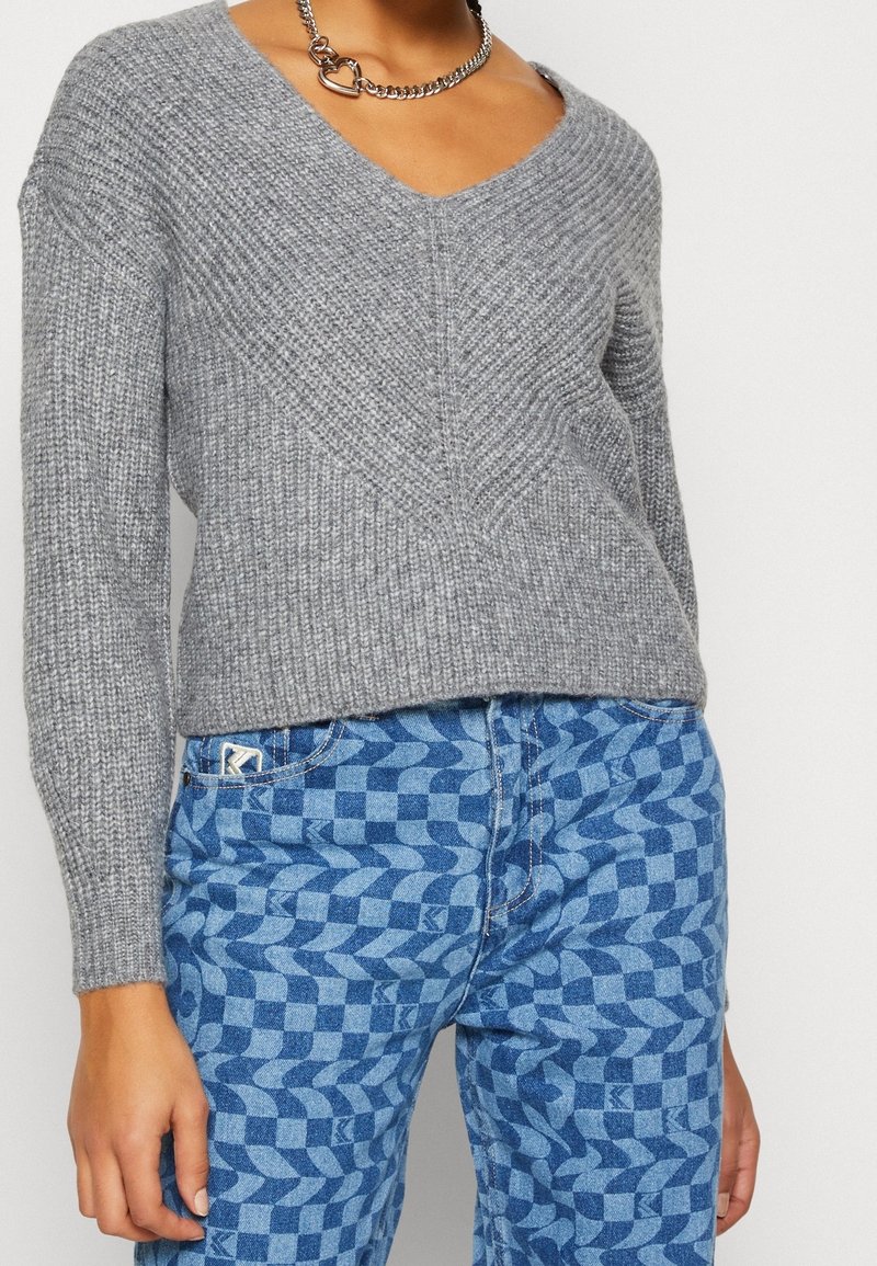 Grauer gestrickter Pullover mit V-Ausschnitt und strukturiertem Rippmuster, kombiniert mit blauen gemusterten Jeans mit Wellenmuster. Silberne Kettenhalskette.