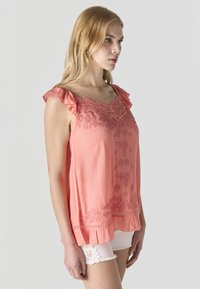 Mujer rubia con blusa sin mangas color coral con detalles de encaje y shorts blancos con borde de encaje, mirando hacia la derecha sobre un fondo liso.