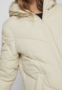 Woman wearing a cream-colored quilted puffer jacket with a hood, front zipper, and visible side pocket, over a black top.