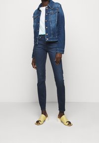 Denim jack in donkerblauw met koperen knopen, gecombineerd met strakke donkere jeans en gele open teen muiltjes met een gebogen band.