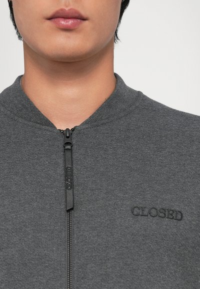 Jachetă gri cu fermoar, fabricată dintr-un material moale, având un guler înalt și un logo negru "CLOSED" pe față, textură netedă și design ajustat.