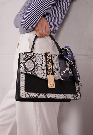 Personne en chemise rayée et pantalon blanc tenant un sac à main à motif serpent, avec chaîne dorée, cadenas et foulard bleu marine en accent.