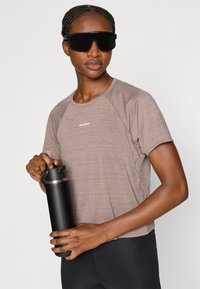 Kortermet, lys brun treningsskjorte med teksturert stoff og logo. Holder en svart vannflaske med sølvlokk, iført store svarte solbriller.